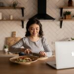 Cheerful ethnic female cutting fresh vegetables on cutting board while sitting at wooden table in kitchen with open portable computer in apartment